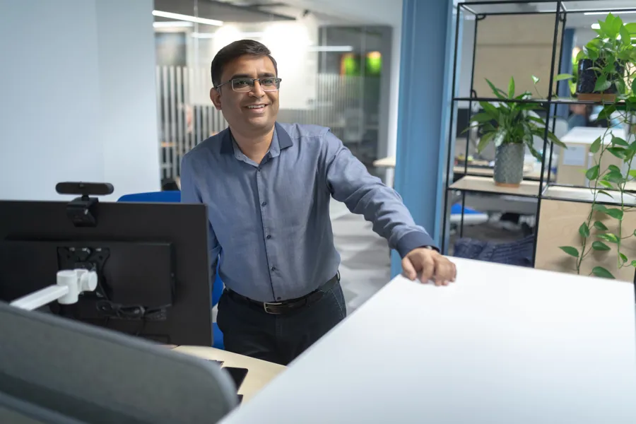 A man in a blue shirt stands at an office desk with plants in the background