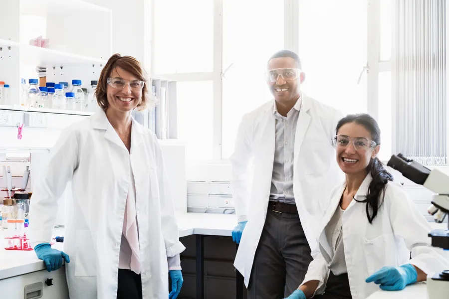 Three scientists in lab coats and safety goggles smiling in a laboratory